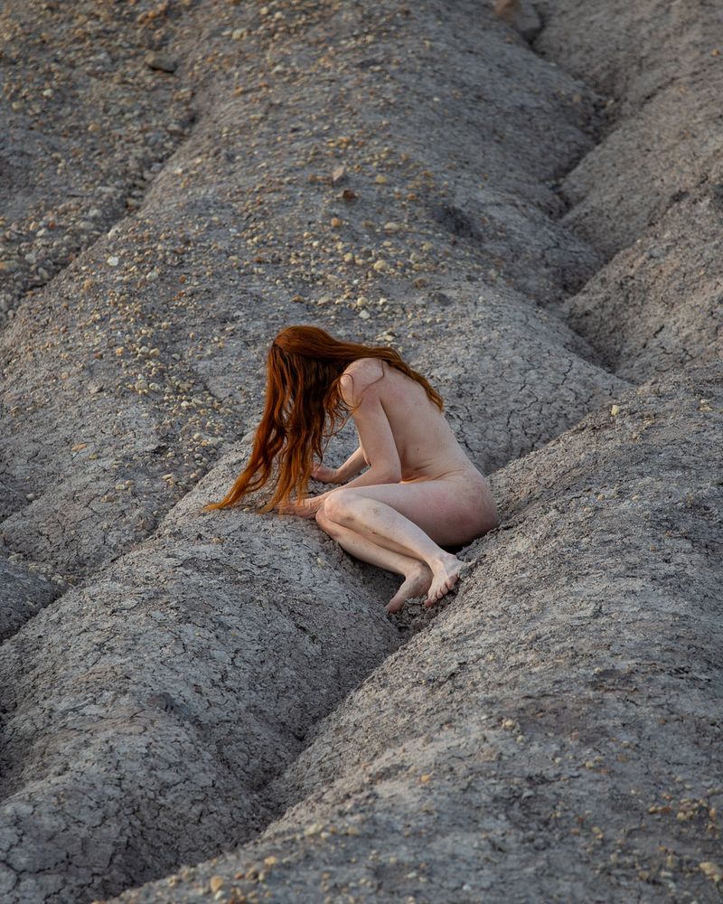 a nude woman with long, fiery red hair is crouching in the slopes of dried out earth at an abandoned open-pit mine.