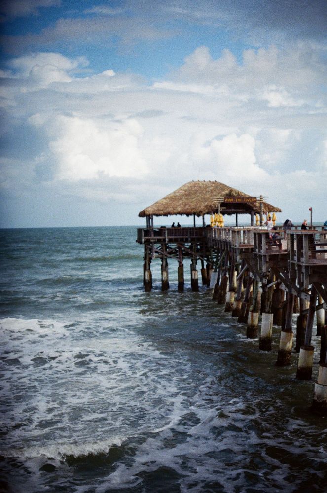 A straw roofed restaurant sits at the end of the pier as fluffy white clouds fill up the top of the screen and the frothy waves warn of rough weather to come
