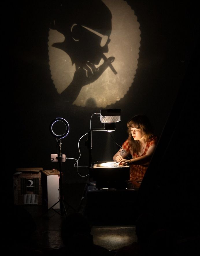 a softened border illuminates a shadow of a woman in glasses with a cigarette as the puppeteer is underlit by the heads up projector being used to create the image.