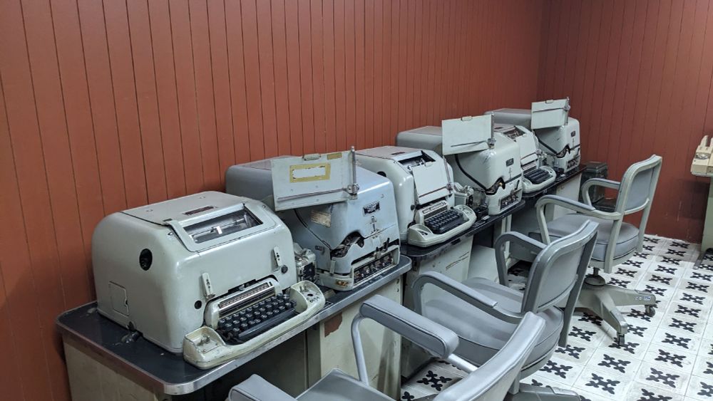 A bank of grey telex machines in an underground room, with grey office chairs and brown walls.