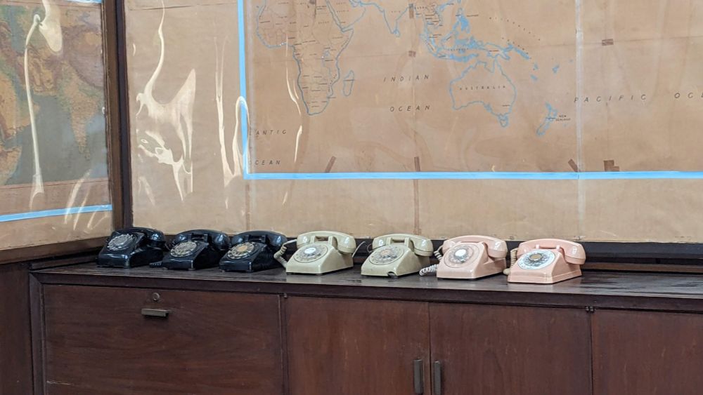 Seven rotary dial phones in colours from black to light pink, close to a world map.