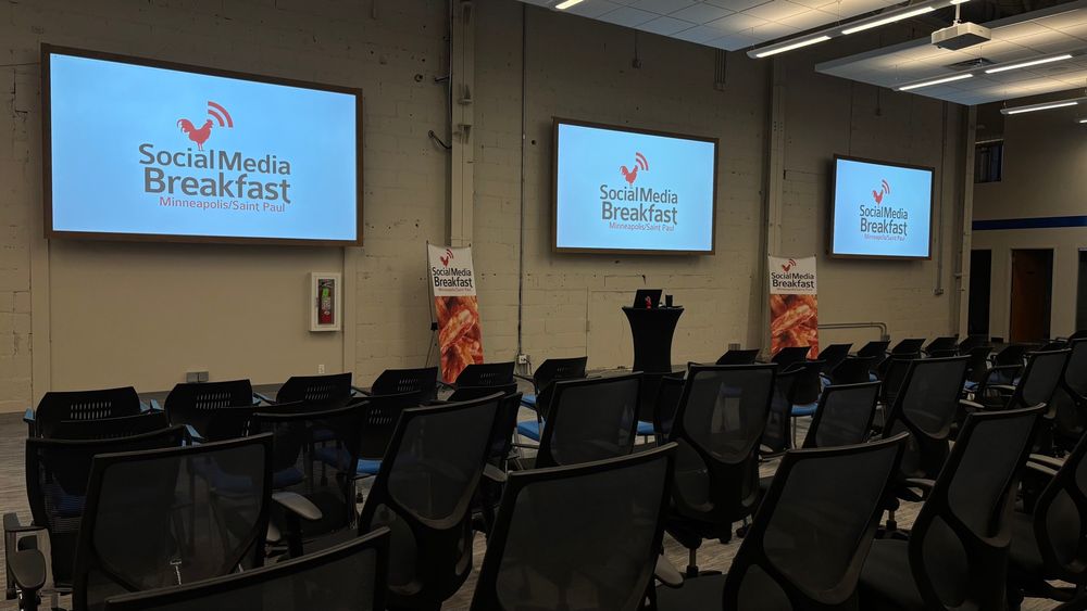 The main space at Improving in Minneapolis. Rows of chairs waiting for butts, and three screens show the Social Media Breakfast logo.