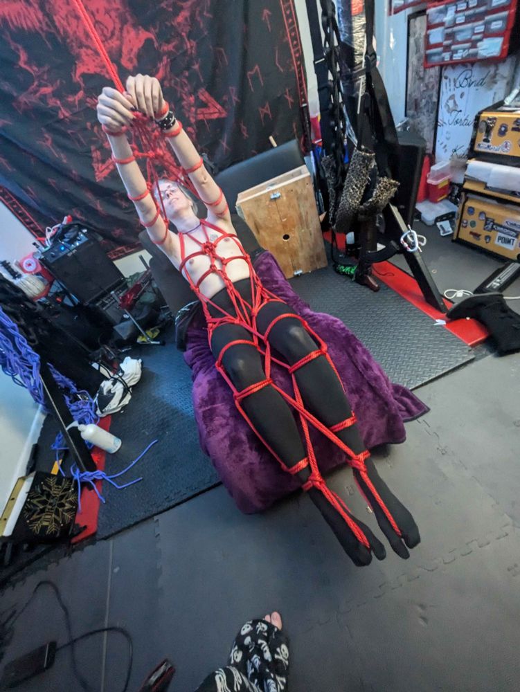 Liv is tied up with red rope, laying on a purple blanket. The rope work is intricate and her hands are bound above her head. 