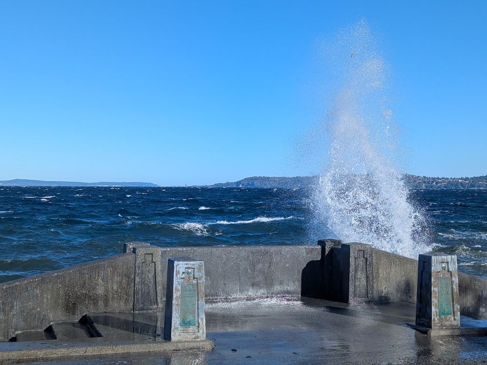 huge surf spray above a concrete seawall barrier, puget sound with rough waters and a blue sky in the background
