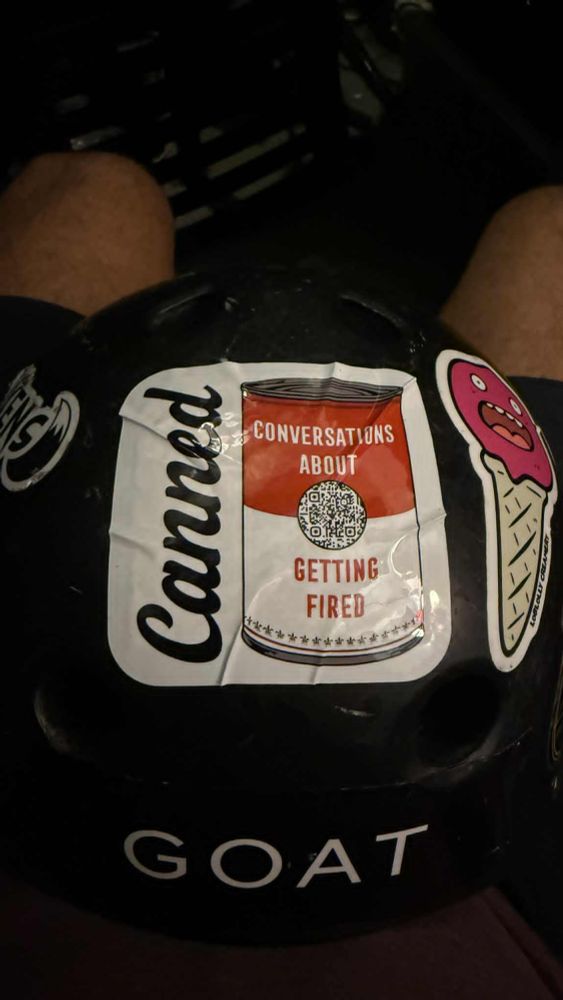 sticker for canned, conversations about getting fired,  on a helmet next to a sticker of a shouting ice  cream cone (you read that right) and above the letters G O A T. sticker shows the canned soup can logo with a QR code. 