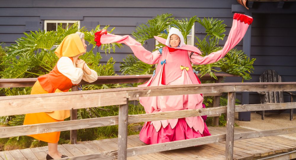 Two cosplayers dressed as the peasant versions of the fairies Flora and Merryweather from Disney's Sleeping Beauty. Merryweather is dressed in the "gown" Flora attempted to make for Princess Aurora. Merryweather's extra-long sleeves are flung into the air with a look of dismay while Flora smiles fondly.