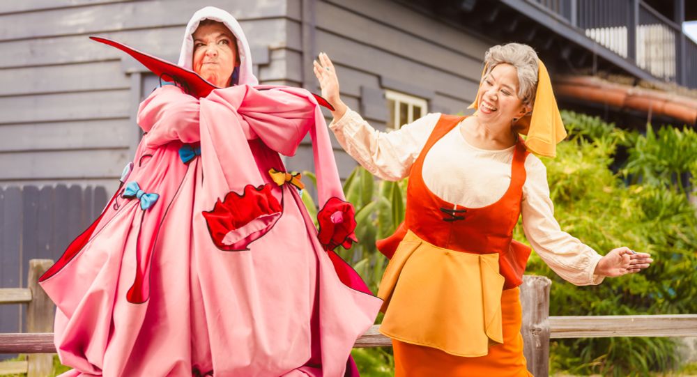 Two cosplayers dressed as the peasant versions of the fairies Flora and Merryweather from Disney's Sleeping Beauty. Merryweather is dressed in the "gown" Flora attempted to make for Princess Aurora. Merryweather has her arm's crossed and looks disgruntled while Flora smiles and appears proud.