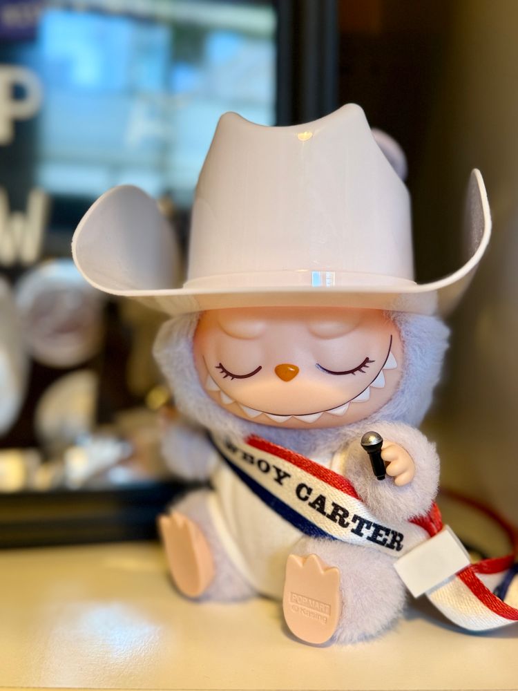 Sitting Labubu holding a LEGO mic with its eyes closed in a cowboy hat and “sash” made from Cowboy Carter tour bracelet given to all attendees. 