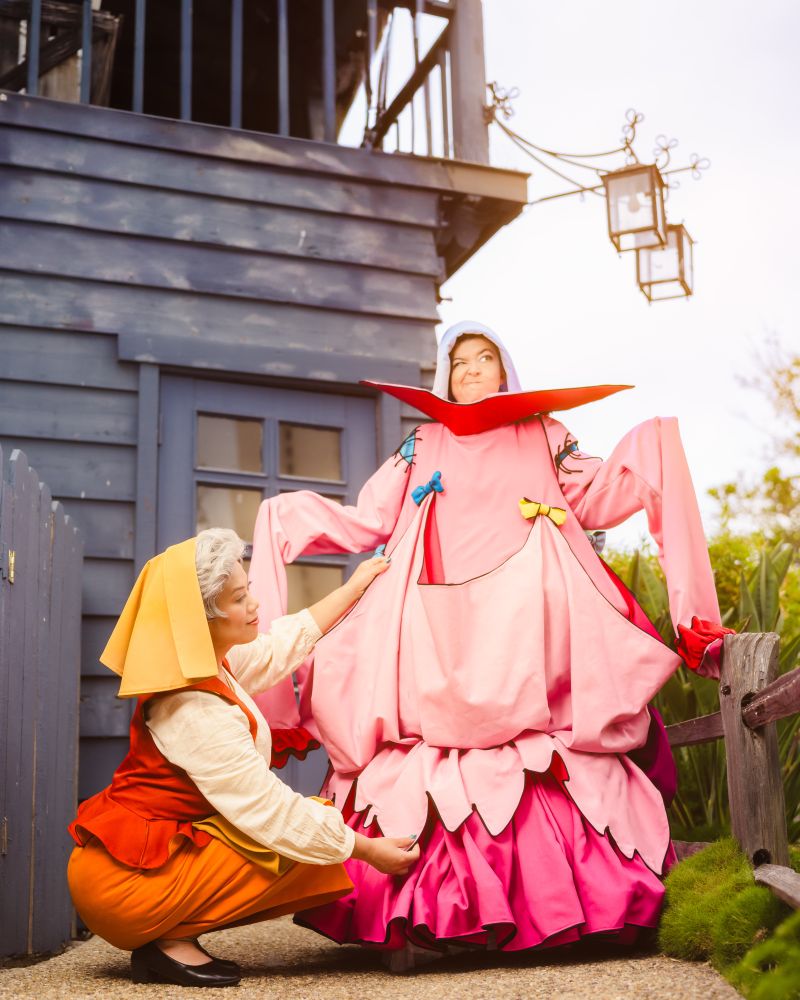 Two cosplayers dressed as the peasant versions of the fairies Flora and Merryweather from Disney's Sleeping Beauty. Merryweather is dressed in the "gown" Flora attempted to make for Princess Aurora.