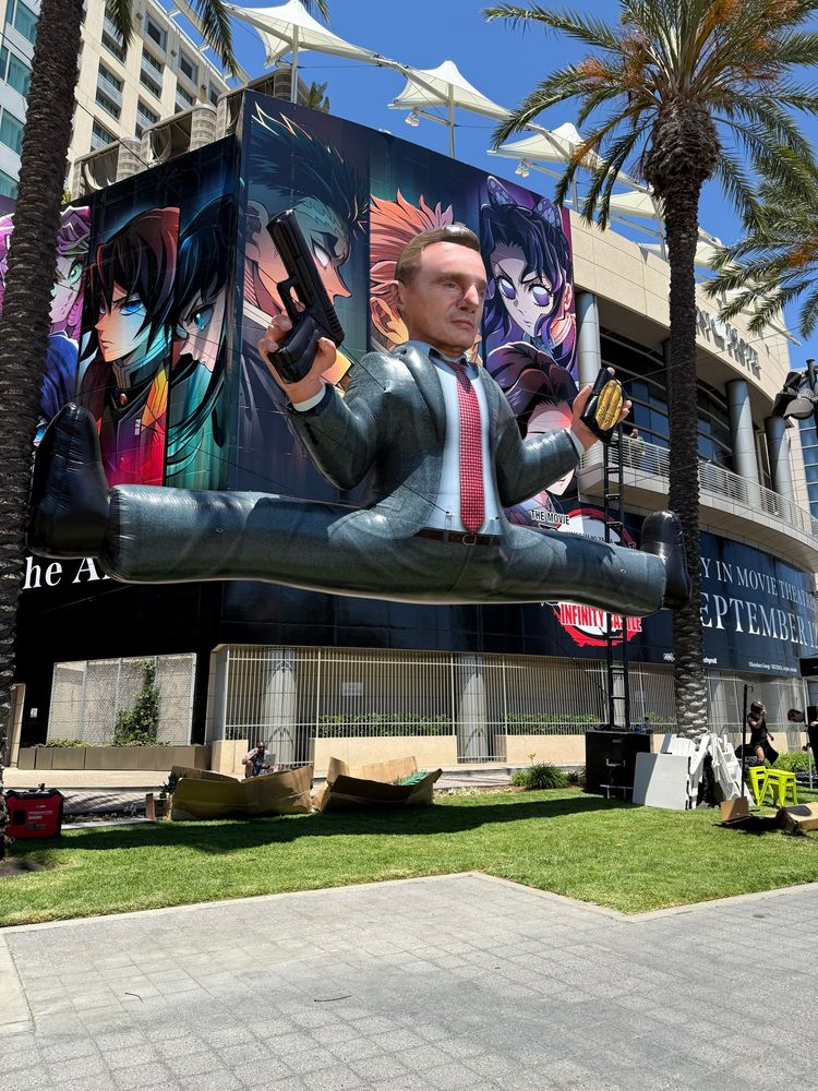 Larger than life inflatable figure of Liam Neeson as Frank Drebin Jr. posed in a split between two palm trees at San Diego Comic-Con. 