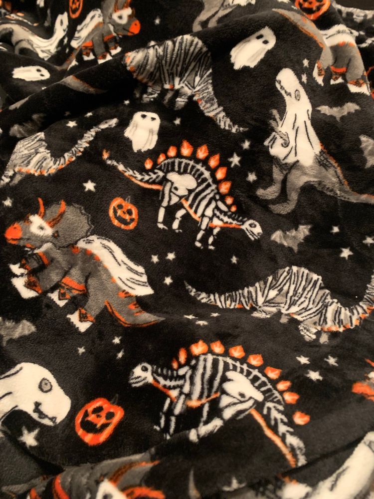 a soft blanket with ghosts, jack-o’-lanterns, bats, clusters of stars, triceratops dressed as superheroes, stegosaurus skeletons, mummified brontosauruses, and t-rexes with sheets over its’ heads that only go down halfway with its arms sticking out of holes on a black background, other colors on it are white and orange 