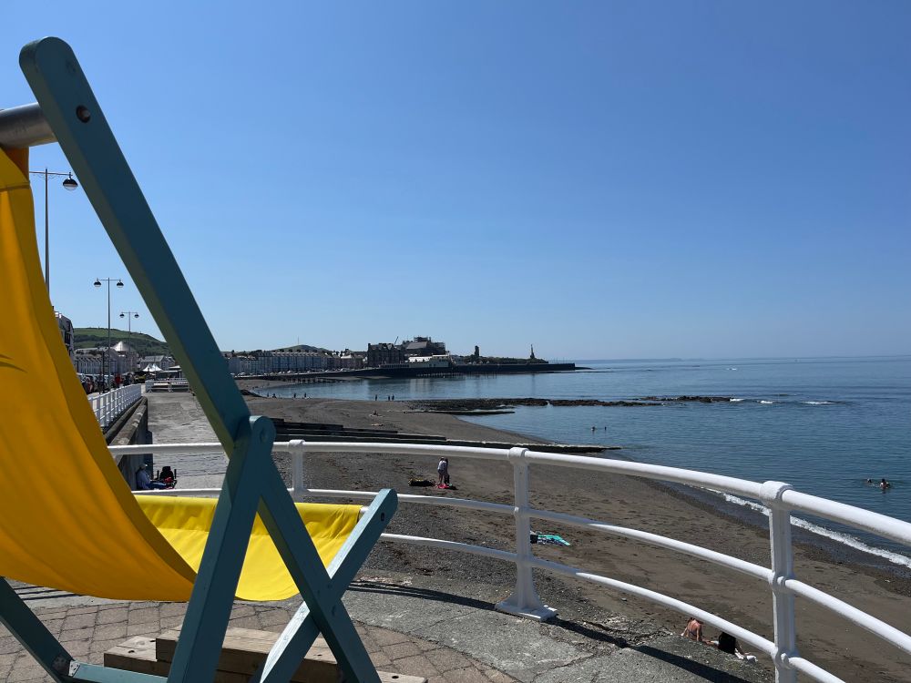 Aberystwyth north beach in the sunshine. A large yellow deckchair is in the foreground and we’re looking from the prom opposite the Glengower over to the pier and castle point. 