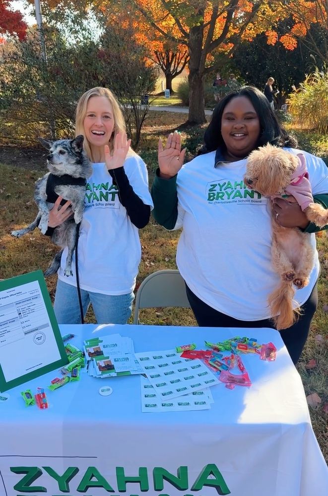 A photo of Adele and Zyahna tabling outside of Charlottesville High School.  Adele is holding Colby, a small gray and black dog, and Zyahna is holding her dog, Zoë, a caramel-colored maltipoo.  Adele and Zyahna are wearing green and white "Zyahna Bryant for Charlottesville School Board" t-shirts, and the table has a matching tablecloth, along with stickers, a sample ballot, and candy. 