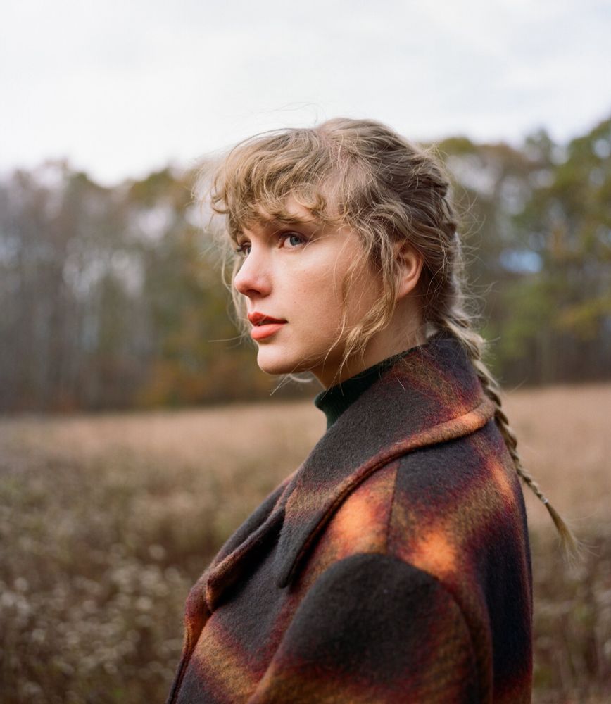 taylor swift wearing a plaid jacket in a field. she has her hair in a braid.
