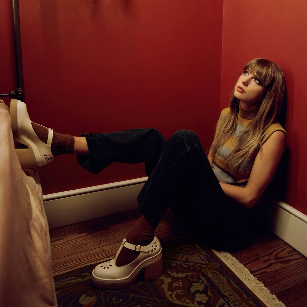 Taylor Swift sitting down with one leg up. She is wearing a striped short sleeve shirt, black pants, maroon socks, & cream mary jane style shoes. shes sitting against a red wall