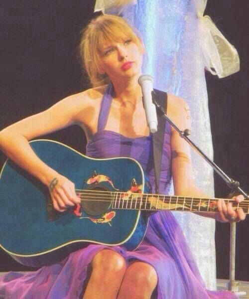 Taylor Swift sitting down in a purple dress. She is holding a blue guitar with koi fish on it & theres a microphone in front of her