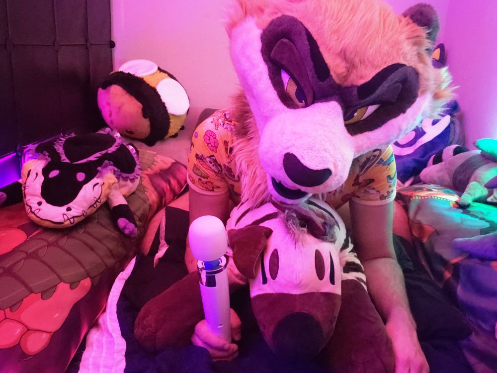 Ferret fursuiter on top on a hyena plush, holding a vibrator with lusty intent, in a cluttered cozy bed