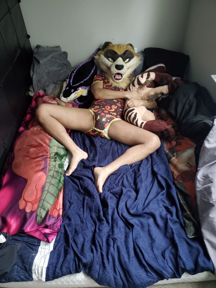 Same bed and fursuiter, only ferret is spread legged while laying down and looking up at viewer, eyelids still on and still holding hyena plush