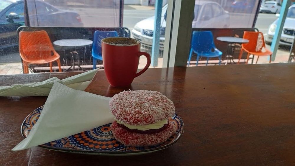 Wooden bench in front of window with red mug of cappucino and pink lamington  sponge kiss. Sunshine outside