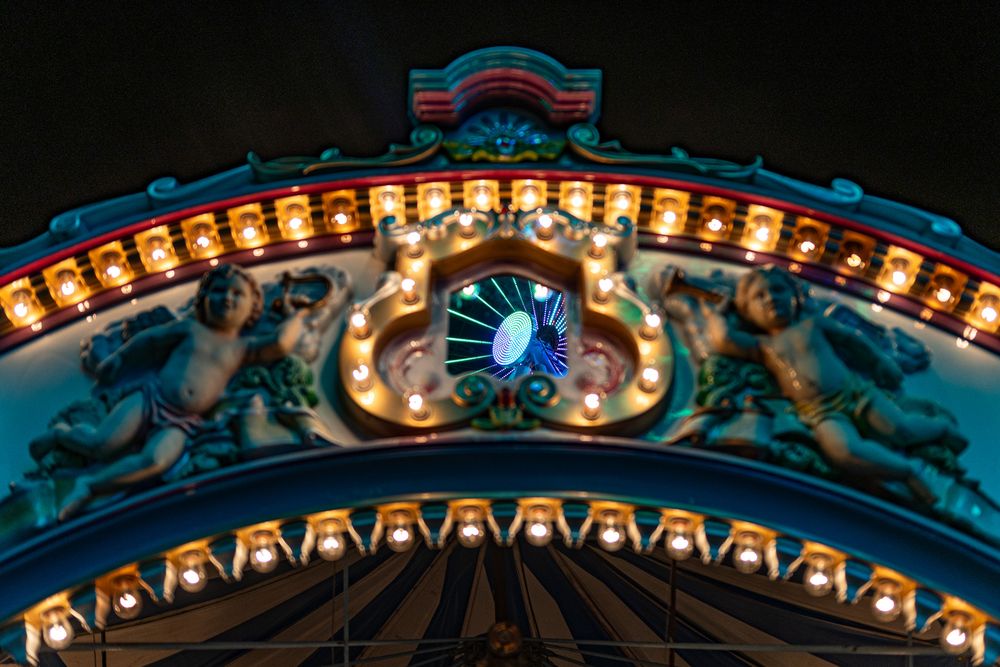 A photograph of a top of a merry-go-round that in the center has a mirror that is reflecting a colorful center of a Ferris Wheel.
