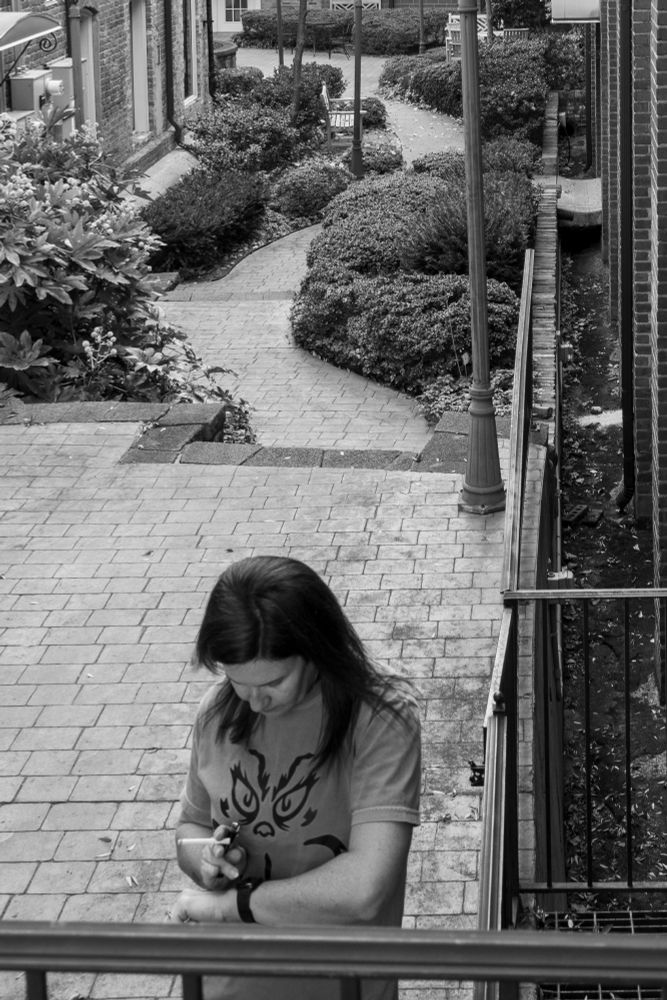 A black and white photograph of a woman with a ciggarette in her hand checking her Apple Watch. Behind her there are stairs and a path through a small courtyard.
