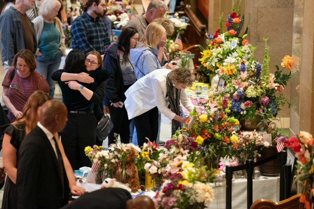 People paying respects and hugging in front of the memorial for the Hortmans outside of the House chamber. Tables are full of flowers and remembrances.