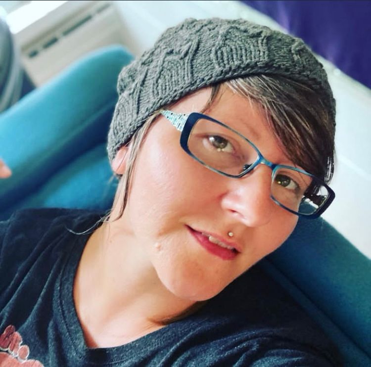 a white woman with a dark grey shirt and knit hat and blue-green rectangular glasses is smiling and leaning her head back on a velvety blue couch.