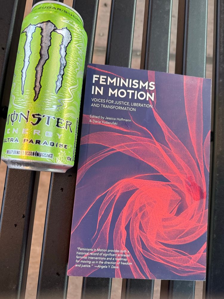 A book (Feminisms in Motion by Jessica Hoffman and Daria Yadacufski) sits on a train station bench beside a can of monster.