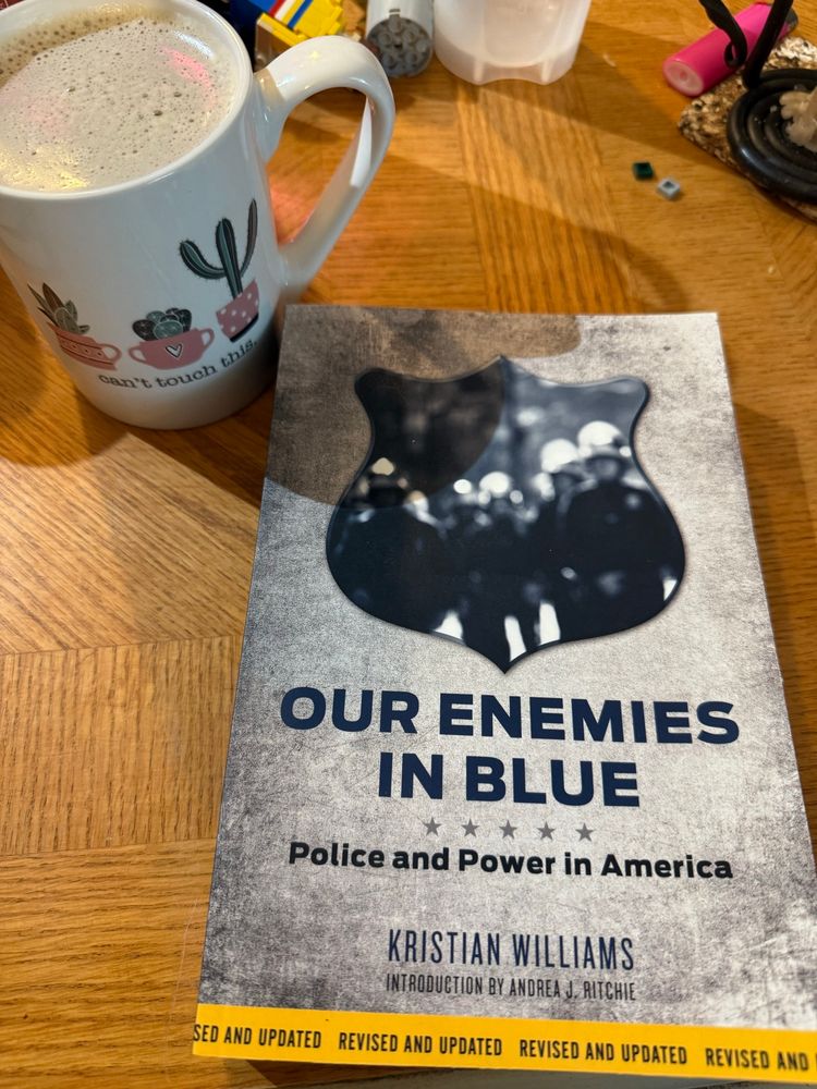 A book (Our Enemies In Blue by Kristian Williams) sits on a coffee table beside a mug of coffee.