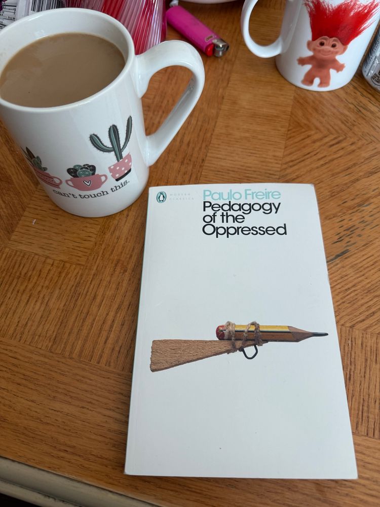 A book (Pedagogy of the Oppressed by Paulo Freire) sits on a coffee table beside a mug of coffee.