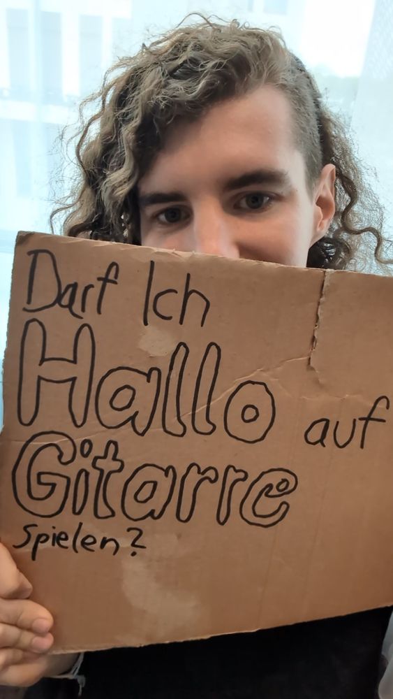 Ich, wir ich ein Schild vor mich halt. Auf dem Schild steht "Darf ich Hallo auf Gitarre spielen"