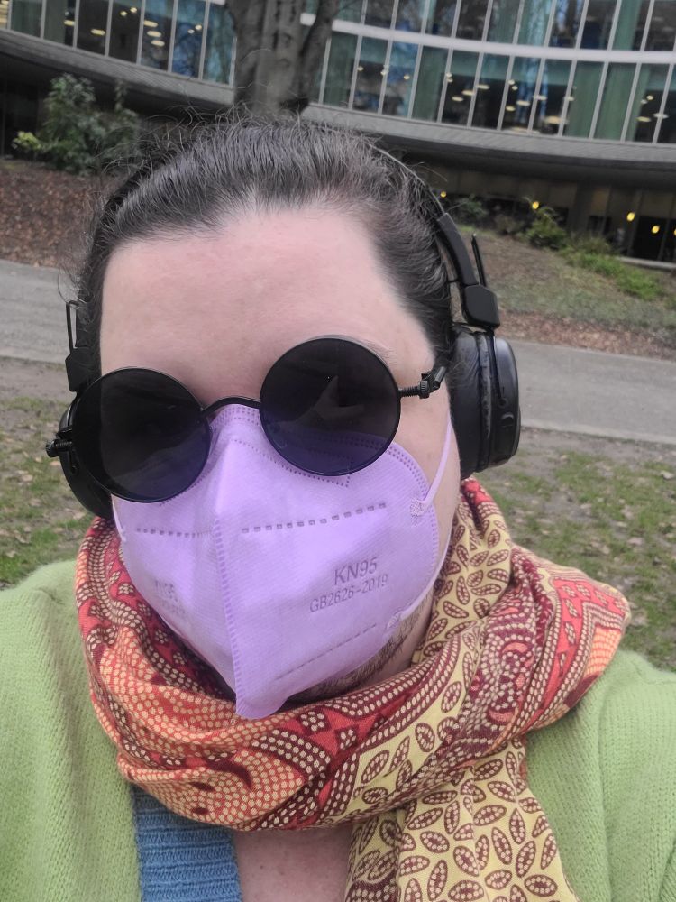 A white nonbinary person who is outside in front of a building with a lot of windows. They have a green sweater on and a yellow and orange scarf. A lavender kn95 mask and small back circular sunglasses. They are wearing headphones.