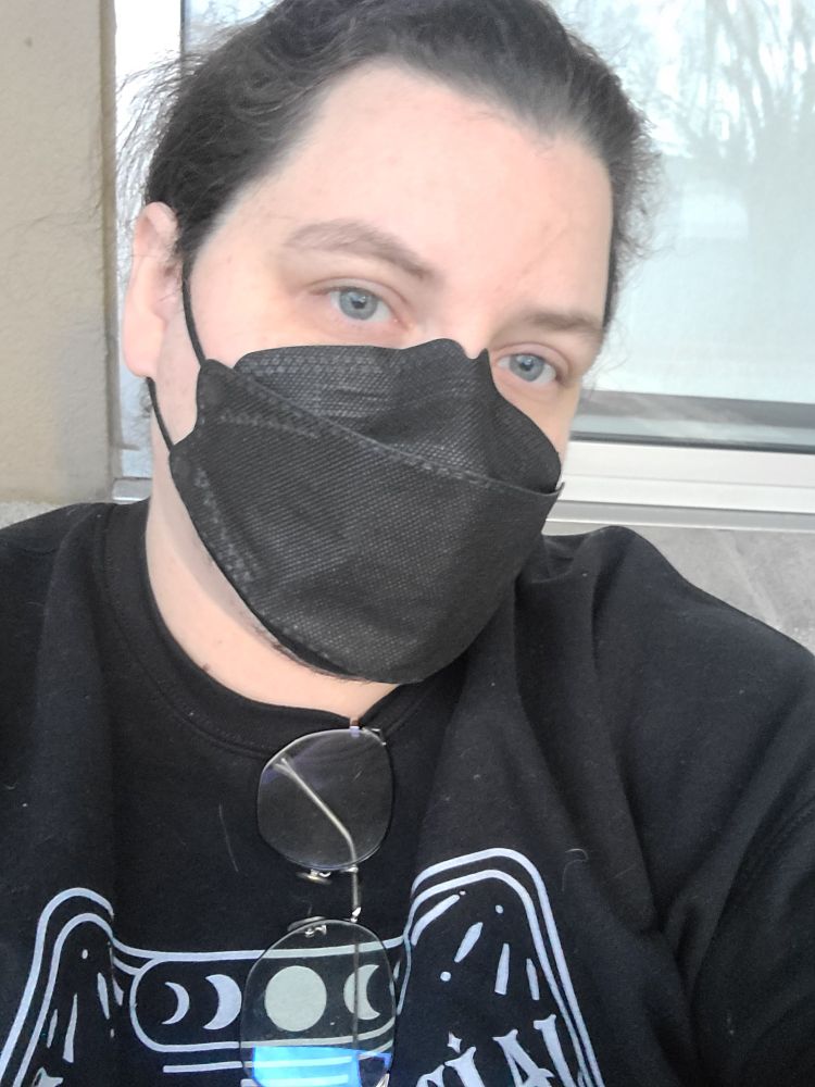 A white nonbinary person who is making eye contact with the camera in a selfie pose. They have on a black face mask and a black sweatshirt. There is a window and a beige wall behind them.