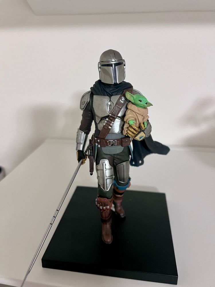 Action figure japonês do Mandalorian com o baby yoda np seu colo e sua lança na outra mão. Sua pose é como se estivesse andando.