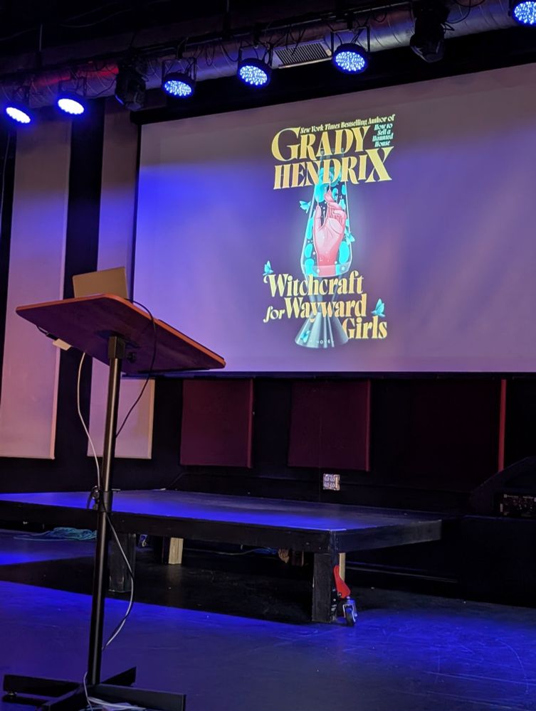 A stage is indirectly lit by soft blue light. Behind a podium, a projector screen displays the cover art for author Grady Hendrix's new book, "Witchcraft for Wayward Girls." The art consists of an ethereal hand rising from a black and turquoise lava lamp. 