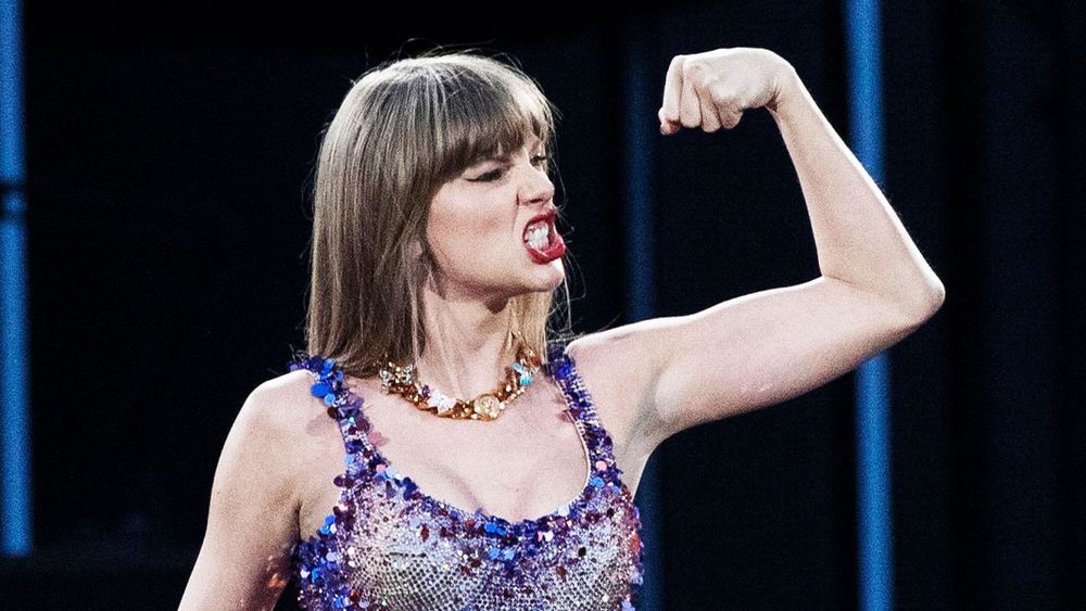 Taylor swift flexing her arm and showing her teeth while wearing a purple lover bodysuit 