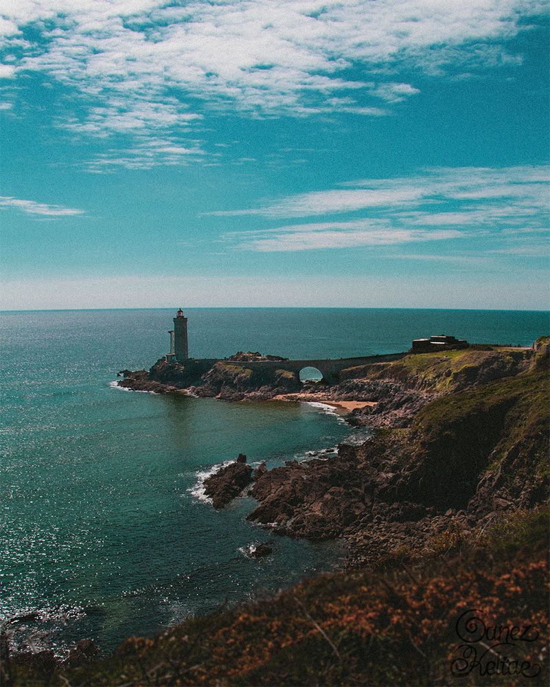 Photo en format portrait d'un paysage marin. Sur le premier plan des plantes à fleurs jaunes sont floues. Au-delà, il y a le phare du Petit Minou, dont la structure est faite de pierres grises et le dôme contenant la lumière est rouge. À sa droite, il y a un pont de pierre construit au dessus de la mer, qui le relie au fort du Minou et à la terre. Diverses plages et rochers habillent la partie droite de la photo, surplombée de parterres herbeux. Enfin, le ciel occupe le tiers haut du cadre. Il est nuageux et d'une couleur similaire à la mer.