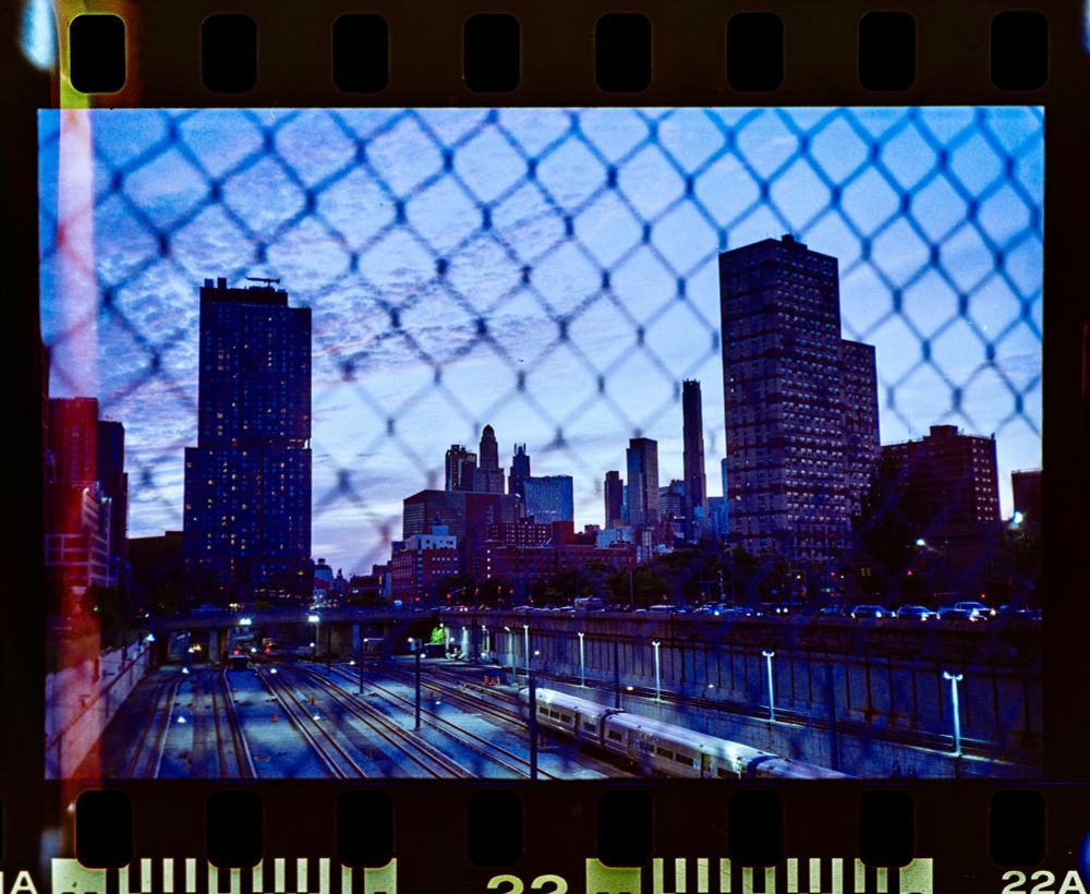 Blueish pink cotton skies over Brooklyn, New York during magic hour. The freshly lit streetlights illuminate the subway tracks below. The scene is framed through a chain link fence and captured by a Canon A35 camera on Kodak ColorPlus 200 film. A slight light leak appears on the left edge. The negative film was scanned and cropped with the sprockets in view. 