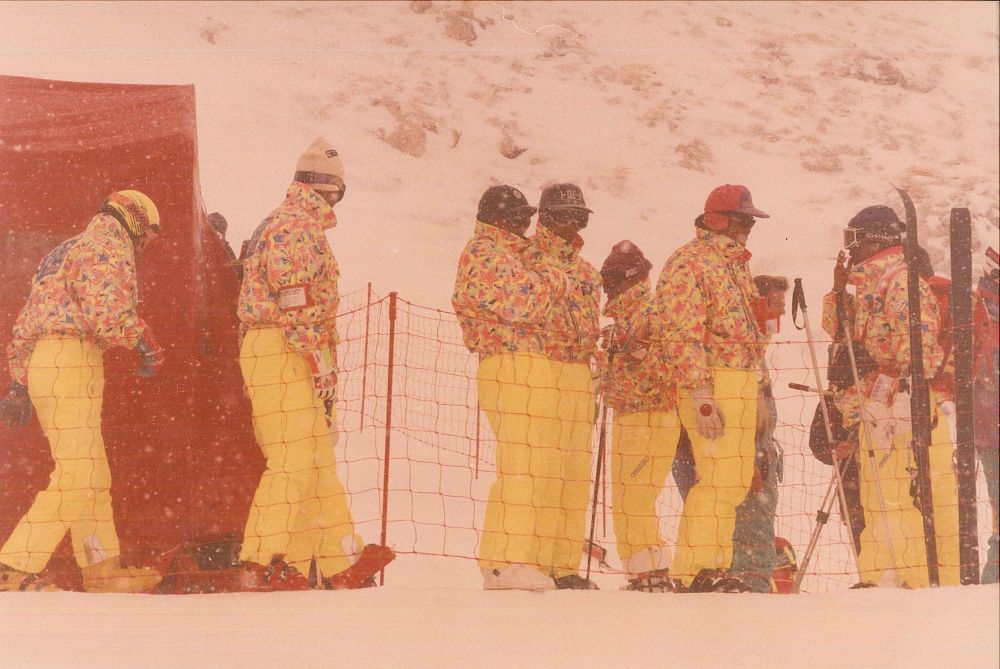 An unknown team relaxing behind a red string fence. Snow is coming down quite heavily, but it's not heavy enough to obscure what they're wearing: hats, ski goggles, patterned jackets that have a series of yellow and pink stars on them, and trousers in such a bright yellow that they could pierce through a snowstorm. One of them is having a smoke.