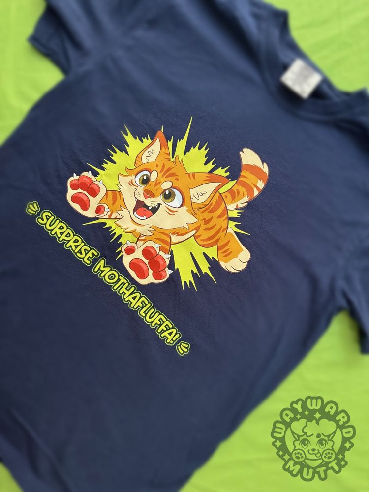 a blue tshirt with an orange kitty jumping forward, text reads "surprise mothafluffa!"