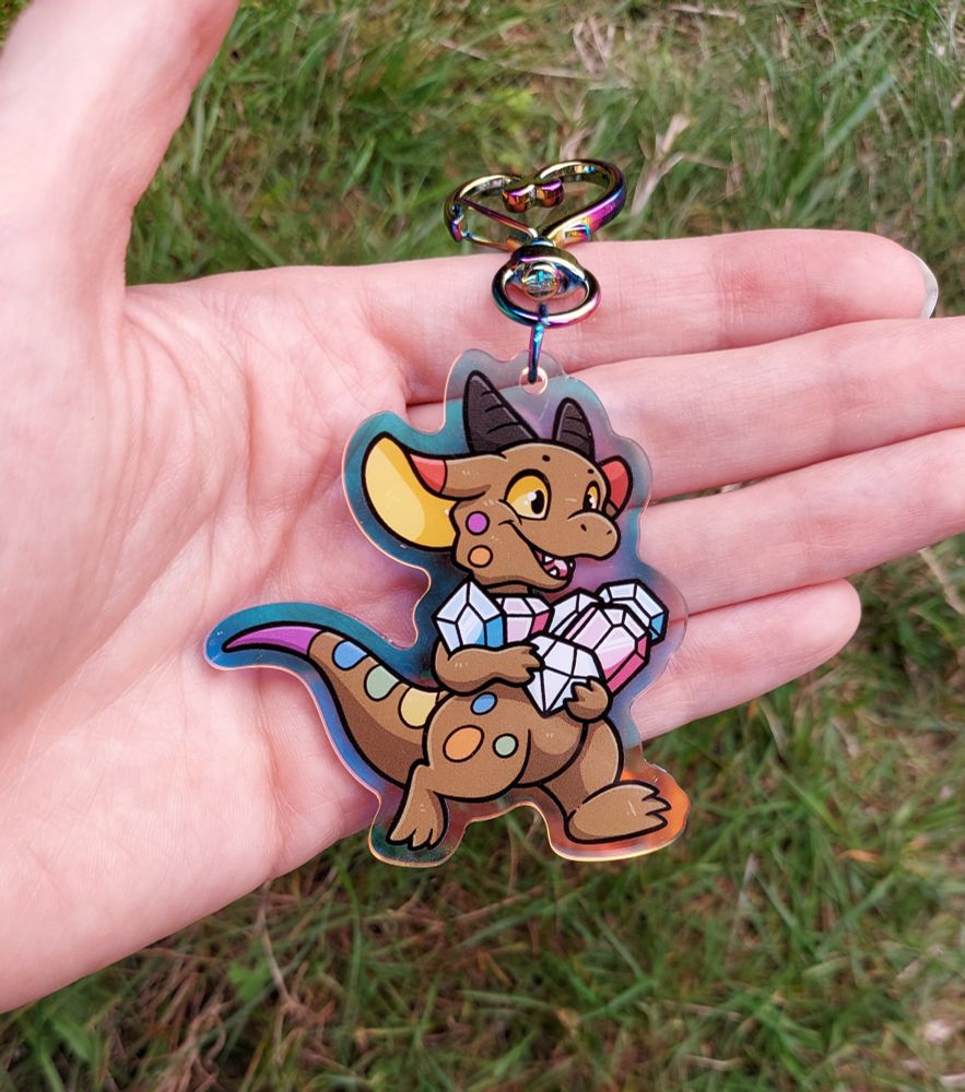 An acrylic charm featuring a kobold in the pride flag colours, running with arms full of gems that are in the trans flag colours
