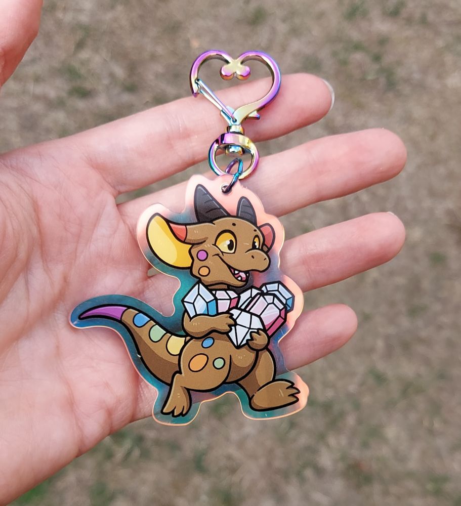 An acrylic charm featuring a kobold in the pride flag colours, running with arms full of gems that are in the trans flag colours. This angle shows the golden reflective hue of the outer edges of the charm.