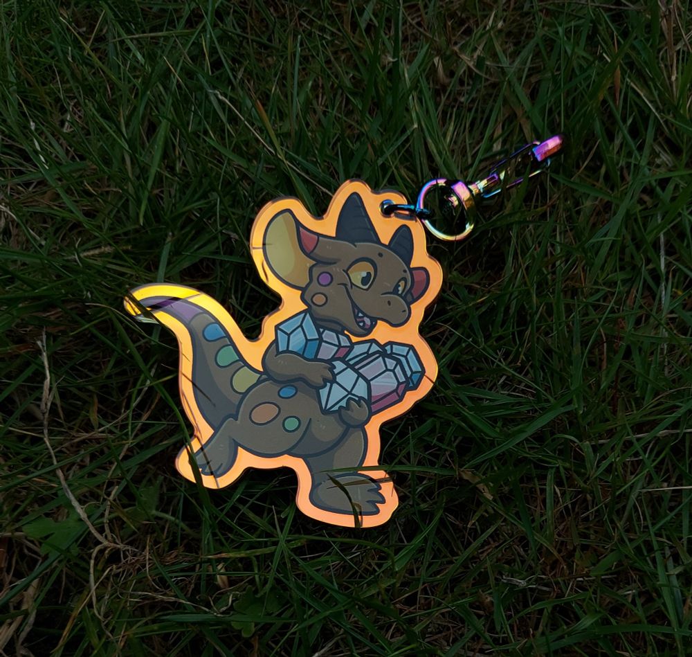 An acrylic charm featuring a kobold in the pride flag colours, running with arms full of gems that are in the trans flag colours. This angle shows the golden reflective hue being very bright in low light.