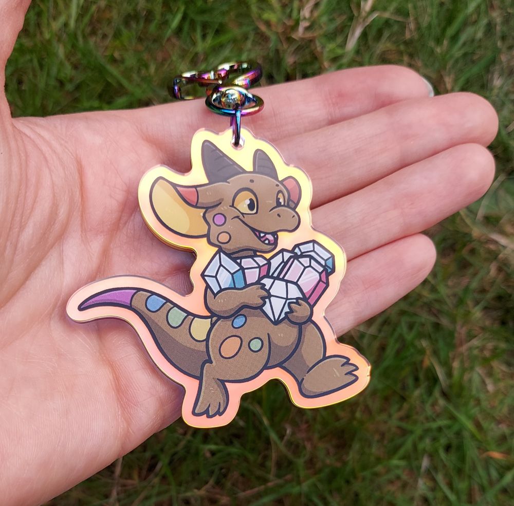An acrylic charm featuring a kobold in the pride flag colours, running with arms full of gems that are in the trans flag colours. This angle shows the golden reflective hue of the outer edges of the charm.