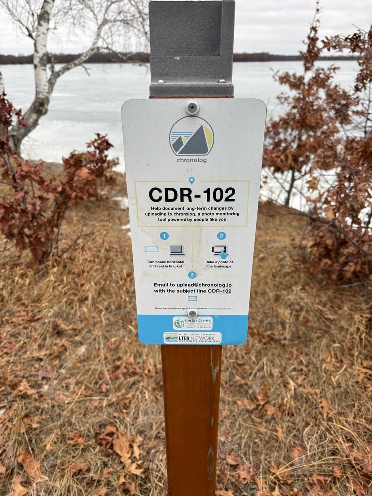 A sign describing how to contribute to long-term photo records with a lake in the background.