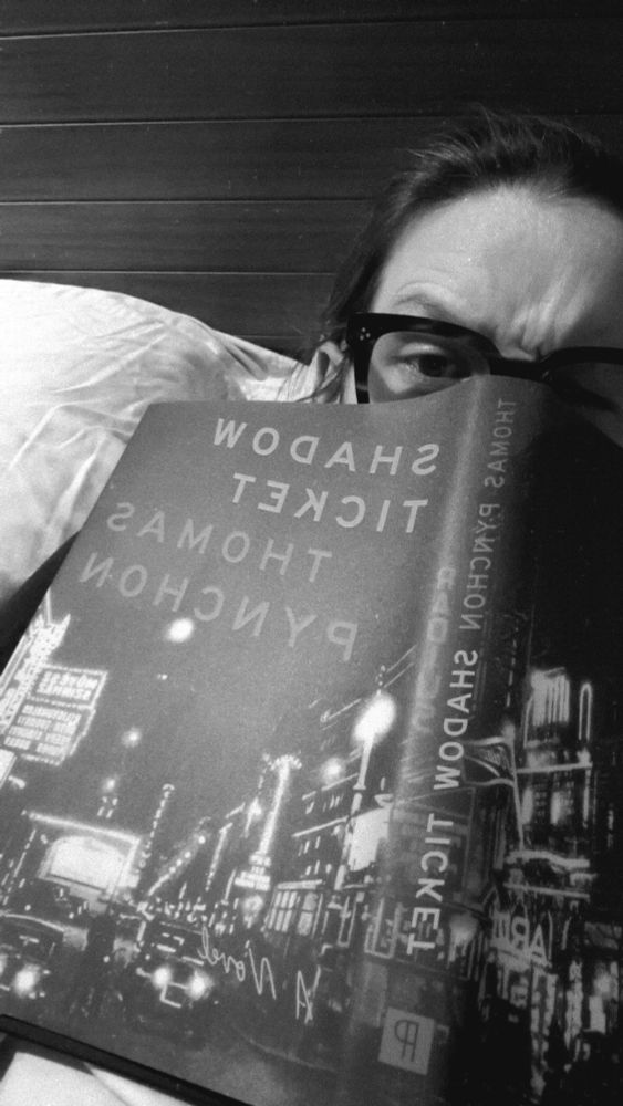 Shadow Ticket by Thomas Pynchon covering the bespectacled face of the reader sitting in bed against pillows and a headboard. 