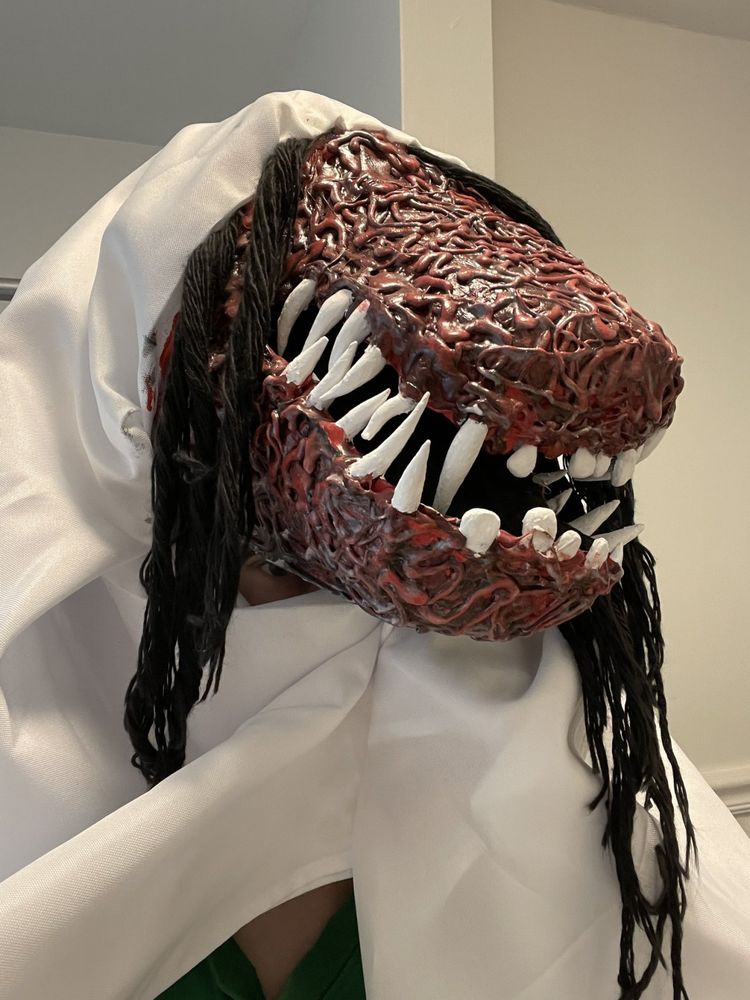 a bizarre mask of a large mouthed monster whose skin texture seems to be made of worms. stringy black hair unfurls from behind a white cloak