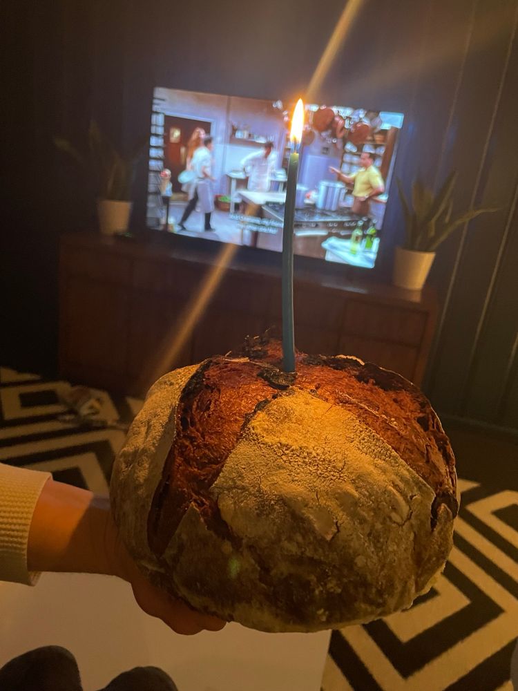 A loaf of chocolate bread with a blue candle burning in it. In the background, Big Night plays on the TV