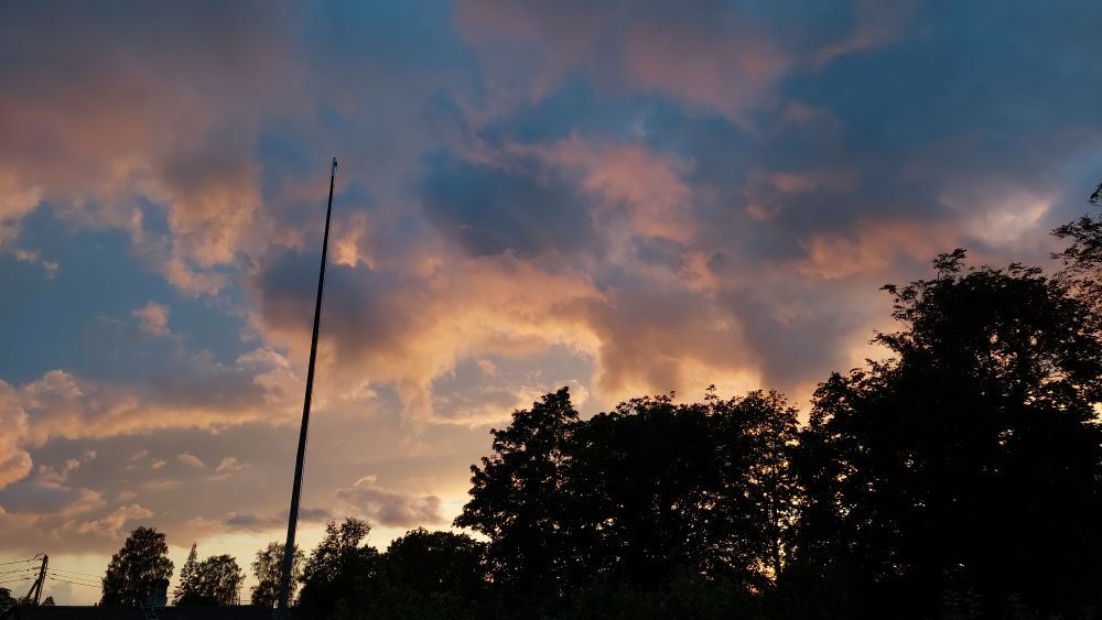 Picture of the sunset, the clouds are a combination of blue and pinkish orangeish. There are trees in the lower right corner and a flag post on the left side of the pic.