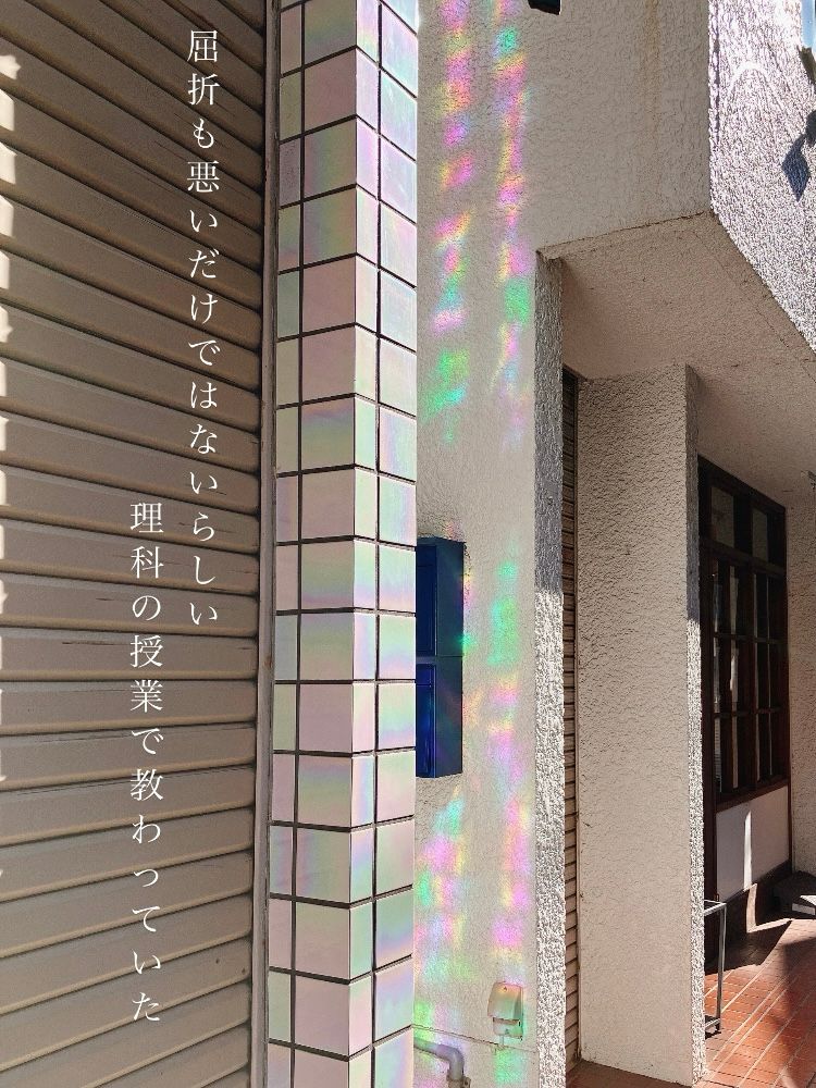 建物の外壁に虹色の反射光が映し出されている写真。左側には縦書きの白文字で「屈折も悪いだけではないらしい  理科の授業で教わっていた」と記されている。